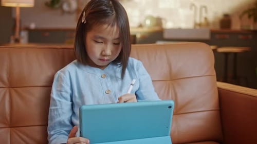 Curious Asian Preschool Kid Girl Using Digital Tablet Technology Device Sitting on Sofa Alone