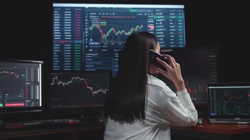 Asian Young Female Stock Market Broker Talking On Mobile Phone With Analysing Graphs