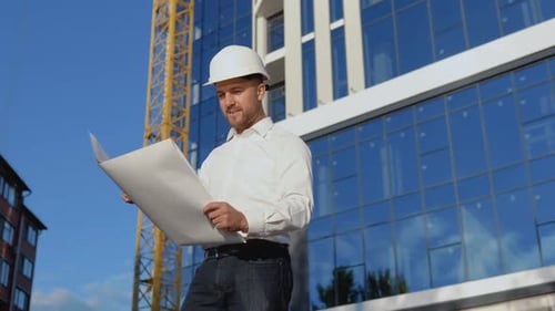 An Engineer in a White Shirt and Helmet Works on the Construction of a Modern Glass Building
