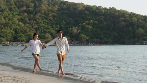 Happy asian couple walking with romantic and holding hand on the beach in summer for leisure.