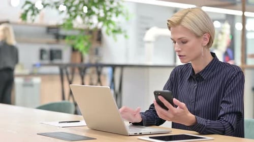 Serious Young Businesswoman Using Smartphone and Laptop in Office