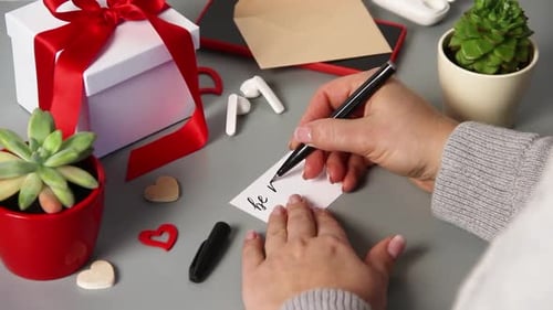 Woman Writing Valentine Card on Desk with Gifts