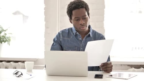Man Reviews Document at Desk with Laptop