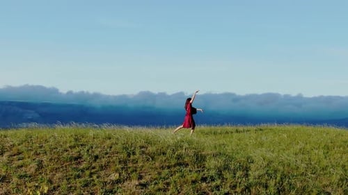 Aerial View of a Young Woman in a Red Dress Dancing on Top of a Hill with a Mountain Range and