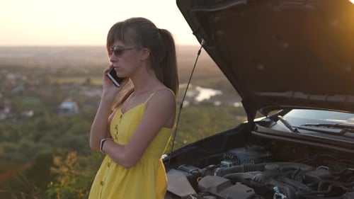 Young female driver talking on mobile phone standing near her car with open hood