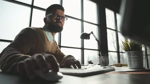 Bearded Man Wearing Glasses Sitting in Office, Working on Computer