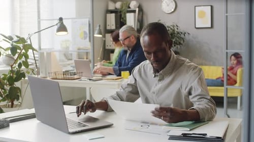 Afro-American Male Office Worker Using Laptop and Reading Document