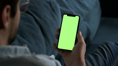 Man lying on couch using smartphone with chroma key green screen at night, scrdoing