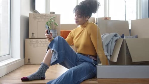 Young Woman Using Mobile Phone In Lounge Of New Home On Moving Day Surrounded By Removal Boxes