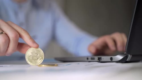 Man Holds Bitcoin at Desk with Laptop