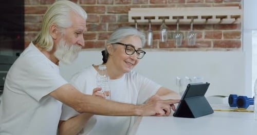 Viejo barbudo y su esposa sentados en la mesa de la cocina y viendo videos interesantes