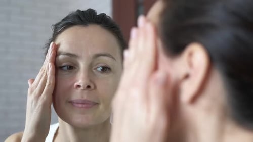 Woman Applying Lotion to Face in Mirror