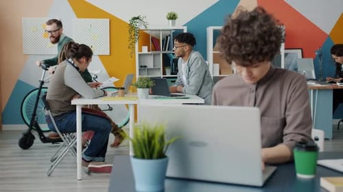 Active Young Man Riding Scooter in Office and Talking To Colleagues Working at Project