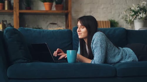 Attractive Young Woman in Casual Clothes Is Typing on Laptop and Smiling While Lying on Sofa in Loft