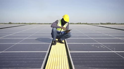 4K African man engineer using digital tablet maintaining solar cell panels on building rooftop.