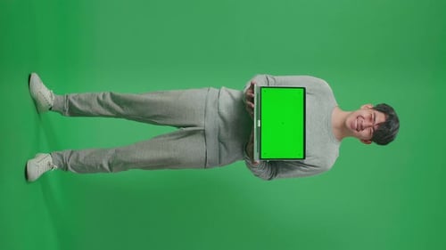 Full Body Of Smiling Asian Man Holding Green Screen Computer While Standing On Green Screen