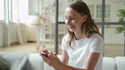 Smiling Woman Uses Smartphone at Home on Couch