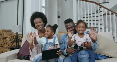 Familia afroamericana con niños sentados en el sofá frente a la cámara y agitando las manos