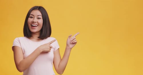 Cheerful Asian Woman Positively Pointing Aside at Copy Space, Orange Studio Background, Mock Up