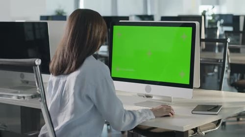 Asian Business Woman Typing on Desktop Computer with Green Mockup Screen While Sitting at His Desk