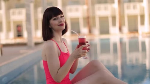 Woman in Elegant Pink Swimwear Relaxing By the Pool in Sunny Day with Refreshing Fruit Cocktail