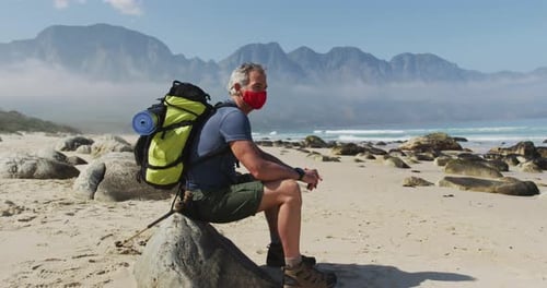 Senior hiker man wearing face mask with backpacks sitting on a rock while hiking on the beach.