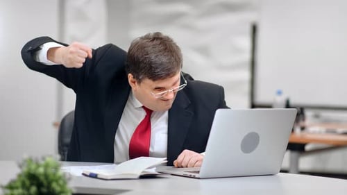 Un patron masculin en colère martèle une table avec fureur en regardant l'écran d'un ordinateur portable, plan moyen sur une caméra rouge