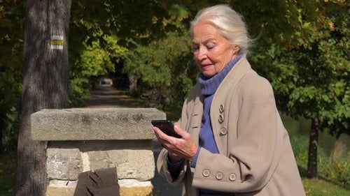 An Elderly Woman Stands on a Bridge and Works on a Smartphone