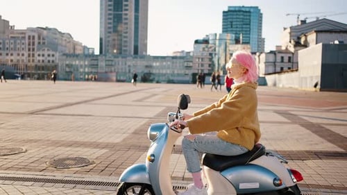 Junge stilvolle Frau mit rosa Haaren fährt auf Roller entlang des Stadtplatzes und genießt gutes Wetter