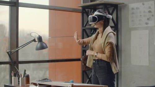 Modern Woman Engineerdesigner in the Office with Large Windows Stands in a Virtual Reality Helmet