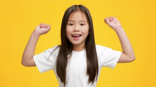 Fröhliches japanisches Mädchen im Vorschulalter schüttelt die Fäuste und feiert Erfolg auf gelbem Hintergrund