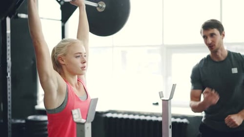 Strong Young Woman Lifting Barbell Focused on Bodybuilding Exercise in Gym with Trainer