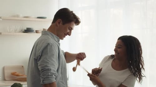 Feliz pareja mixta peleando con cucharas cocinando divirtiéndose en interiores