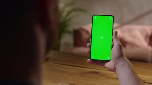 Handheld Camera Back View of Young Man at Home Sitting on a Wood Desk Using With Green Mockup Screen