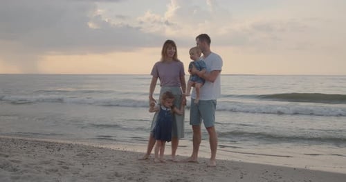 Young Family of Four Stand on the Sea Coast, Holding Their Little Children