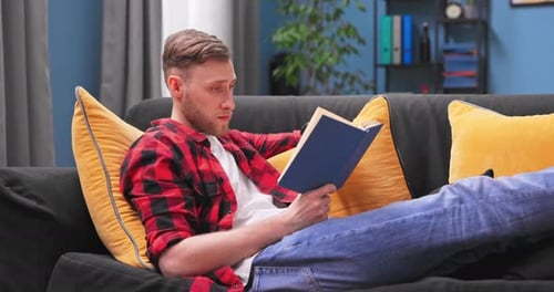 Happy Millennial Boy Lay Relax on Couch at Home Reading Interesting Book Calm Smiling American