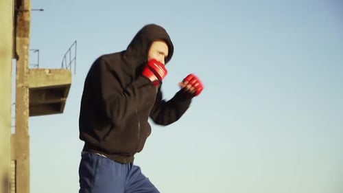 Caucasian Man Boxer in Red Gloves in a Black Sweatshirt with a Hood Makes an Exercise Shadow Fight