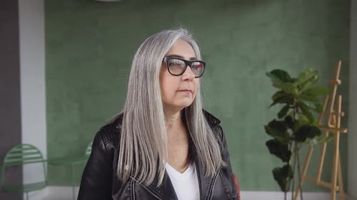 60-Aged Modern Woman in Glasses with Long Gray Hair which Posing on Camera