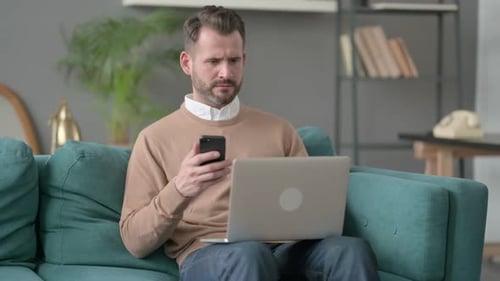 Man Using Phone With Laptop On Couch