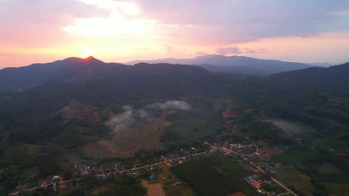 Luftaufnahme einer Drohne, die bei Sonnenaufgang über eine ländliche Gegend mit Bergen im Hintergrund fliegt.