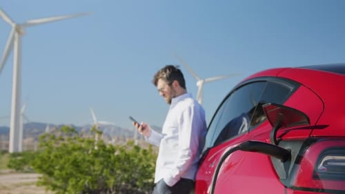 Man Using Phone as Electric Car Charges at Wind Farm