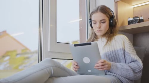 Young woman at home by the window with a tablet in her hands plays, communicates