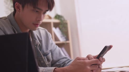 Asian man sits on sofa at home, texting and talking with buddy, holding smartphone,or checking mobil