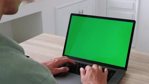 Over the Shoulder Shot of Young Man Sitting at Table and Working From Home on a Laptop Computer with