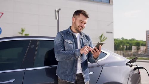 Bärtiger Mann benutzt sein Telefon, während er sein luxuriöses Elektromobil in der Ladestation auflädt