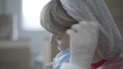 Side View of Young Caucasian Woman in Protective Gloves and Hat Putting on Face Mask. Close-up of
