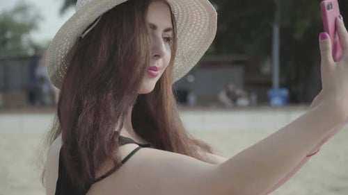 Portrait of Young Woman in the Summer White Hat Taking Selfie on Her Cellphone Resting on the Beach