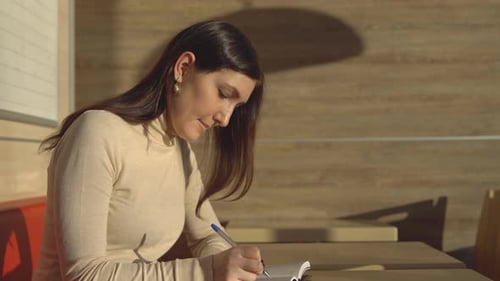 Young Brunette Woman Sitting in a Cafe and Making Notes in a Notebook
