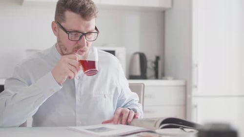 Ein junger stilvoller Mann mit Brille liest morgens beim Frühstück eine Zeitschrift und trinkt Tee