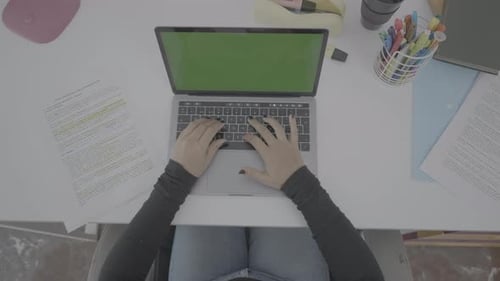 A Young Woman Dressed in Black is Sitting at a Clean Desk Watching a Laptop with a Green Screen
